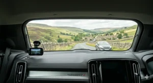 An interior perspective looking out through the rear windscreen of a vehicle, showing a small rear-facing dash cam mounted on the glass, monitoring a grey car following behind on a rural UK road.